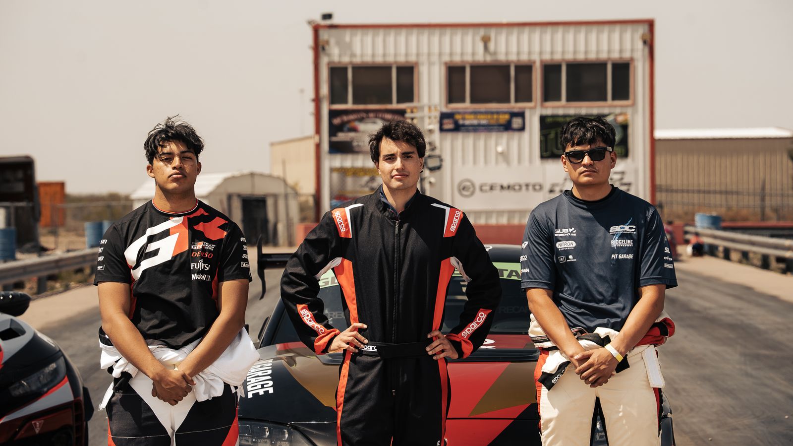 Three team members in race suits at the track, race cars behind them